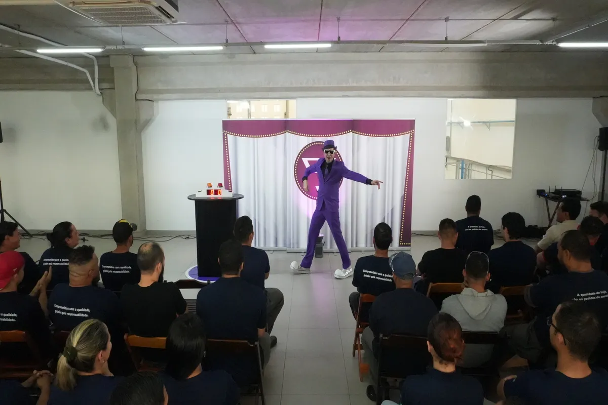 Um palestrante em terno roxo, óculos de sol e chapéu, se apresenta de forma teatral em um palco para um público sentado, durante uma palestra sobre a NR 13.