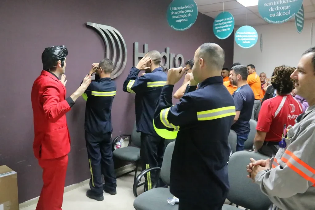 Palestrante de terno vermelho conduz dinâmica com funcionários da Hydro vestidos de uniforme, focada em segurança e uso de capacetes.