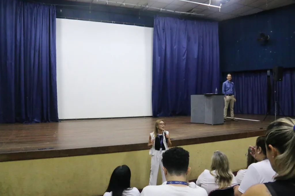 Palestrante de roupa branca falando em palco de auditório, com um homem ao fundo e público em primeiro plano, durante um evento de treinamento sobre NR 5 (CIPA).