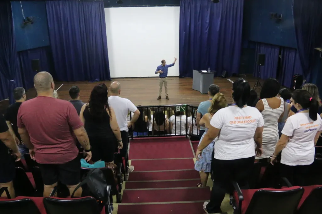 Vista de cima do auditório mostrando o palestrante no palco gesticulando para a plateia que está em pé nas escadas, durante um treinamento sobre NR 5.