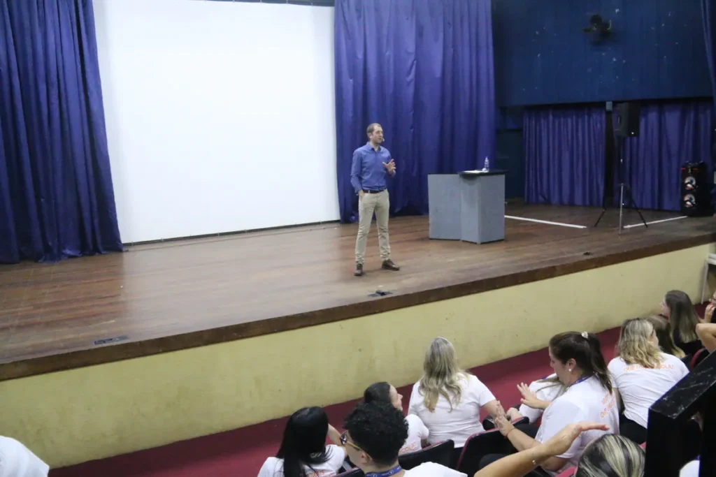 Palestrante masculino de camisa azul e calça cáqui em pé no centro do palco de auditório, preparando-se para falar ou fazendo uma pausa, com a tela de projeção branca ao fundo.