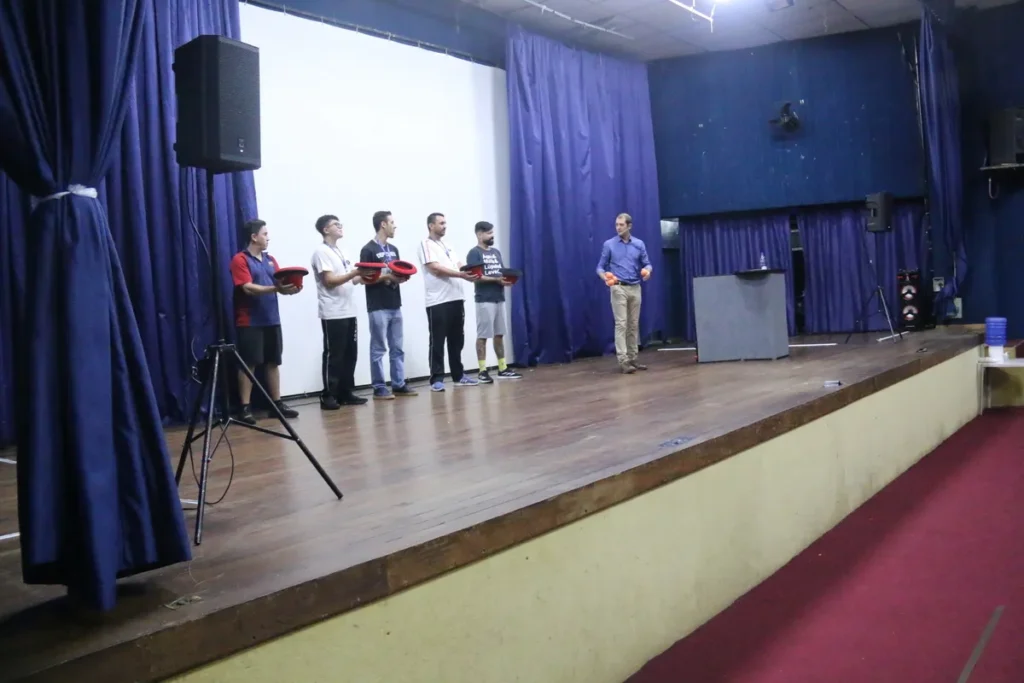 Seis homens em um palco de auditório, cinco segurando objetos vermelhos, ao lado do palestrante, indicando cerimônia de formatura ou entrega de kits de treinamento da NR 5.