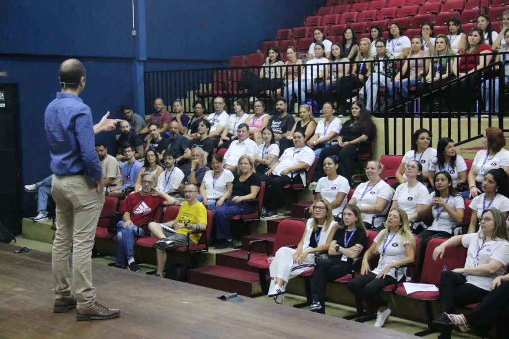 Palestrante de camisa azul em pé no palco de um auditório, gesticulando para a plateia lotada de pessoas em cadeiras vermelhas, durante um evento de treinamento sobre NR 5.