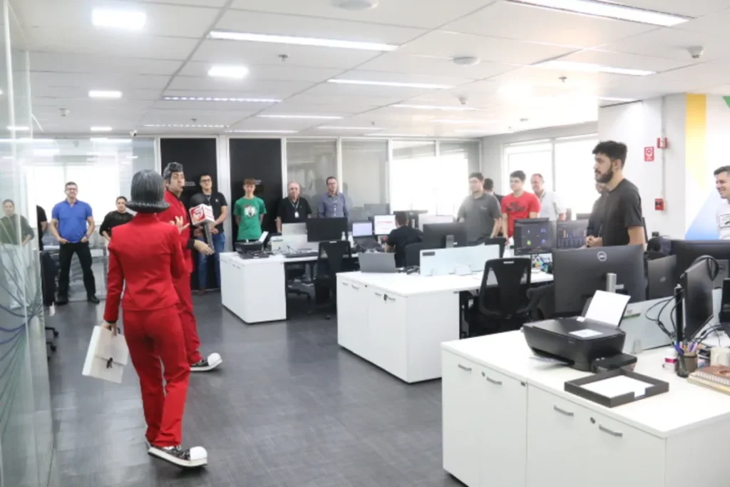 Vista de costas de dois apresentadores em macacões vermelhos, segurando microfones, em um escritório de plano aberto, com funcionários reunidos ao redor para um evento.