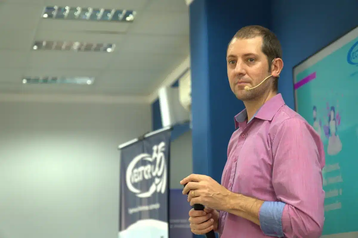 Homem de camisa rosa realizando palestra em evento corporativo com microfone headset e apresentação em telão ao fundo.