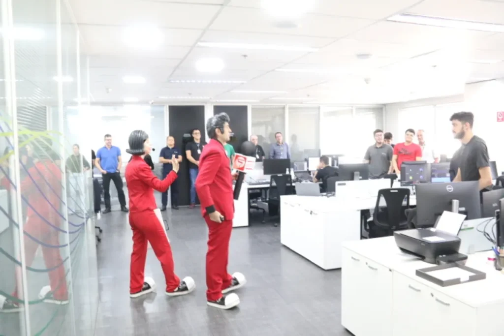 Atores caracterizados em esquetes interativas realizam palestra sobre segurança com objetos cortantes em escritório corporativo, com participação de colaboradores.
