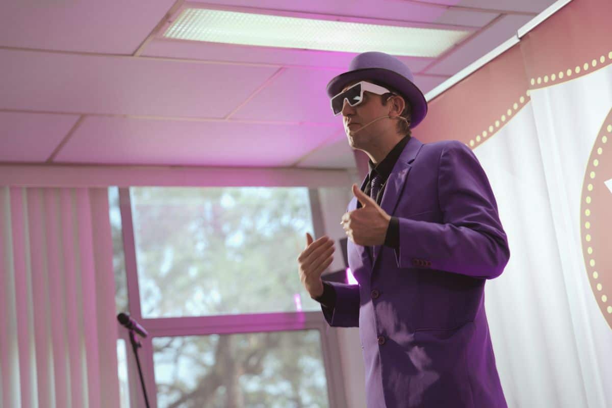 Palestrante com terno roxo, chapéu e óculos conduzindo apresentação ao vivo para evento de SIPAT.