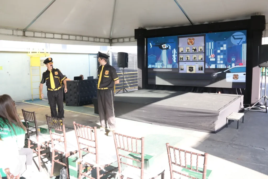 Dois homens vestidos com uniformes temáticos de segurança, com camisas pretas e amarelas e quepe, estão no palco de um evento corporativo realizado ao ar livre sob uma tenda. Eles se preparam para dar uma palestra ou treinamento. Ao fundo, um grande telão de LED exibe gráficos e ilustrações com o tema de prevenção de acidentes e segurança patrimonial, com elementos gráficos de edifícios e veículos. Cadeiras marrons vazias estão dispostas para a plateia.