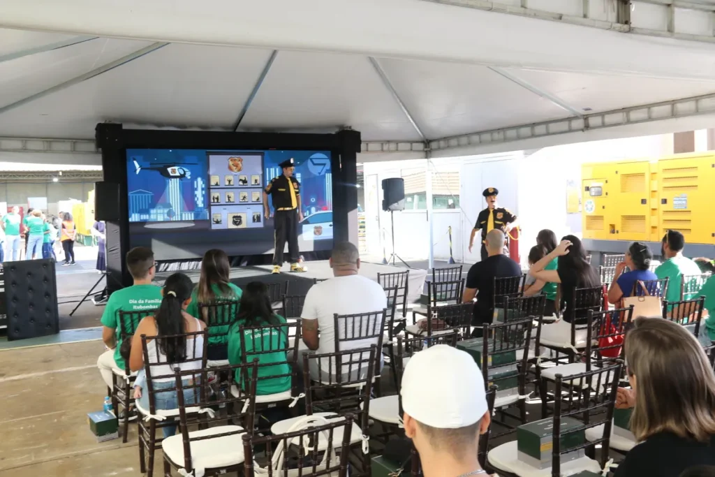 Dois homens vestidos como agentes de segurança realizam uma palestra em um evento corporativo ao ar livre sob uma tenda. O palestrante principal está em frente a um grande telão que exibe ilustrações de segurança e prevenção. A plateia está sentada em cadeiras brancas e de madeira, com alguns participantes usando camisetas verdes. O evento, com tema de Segurança no Trabalho, tem máquinas industriais amarelas visíveis ao fundo, à direita.