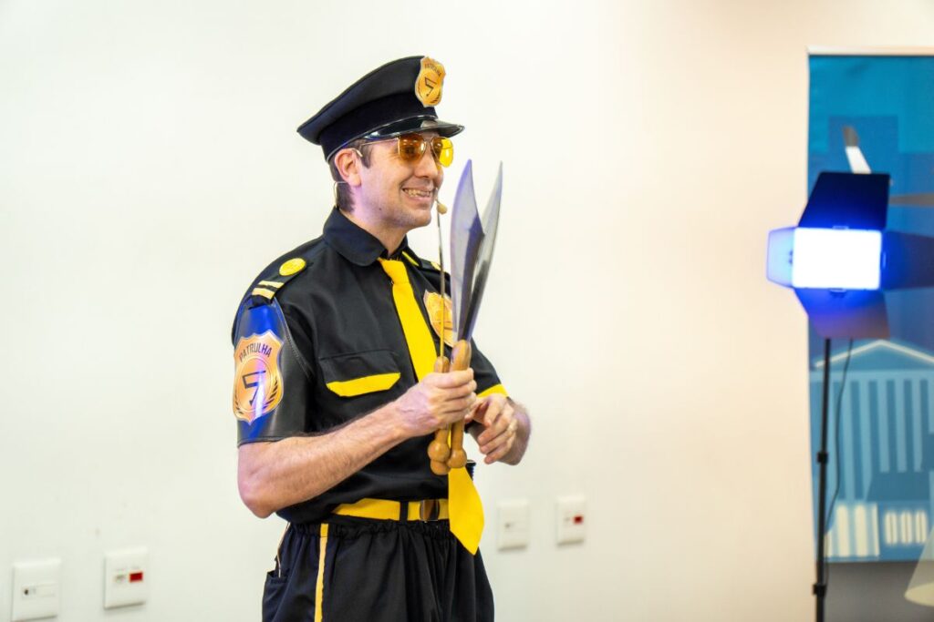 Palestrante vestido como agente de segurança, usando microfone headset e segurando objetos durante apresentação de SIPAT.