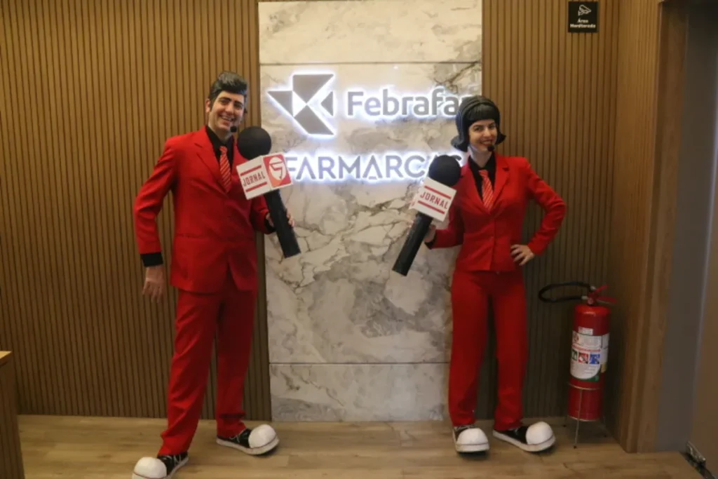 Personagens fantasiados com ternos vermelhos e microfones cenográficos em frente ao letreiro da Febrafar e Farmarcas