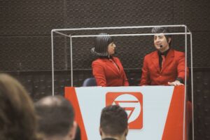 Dois apresentadores com roupas vermelhas participando de apresentação teatral em cenário de SIPAT online.