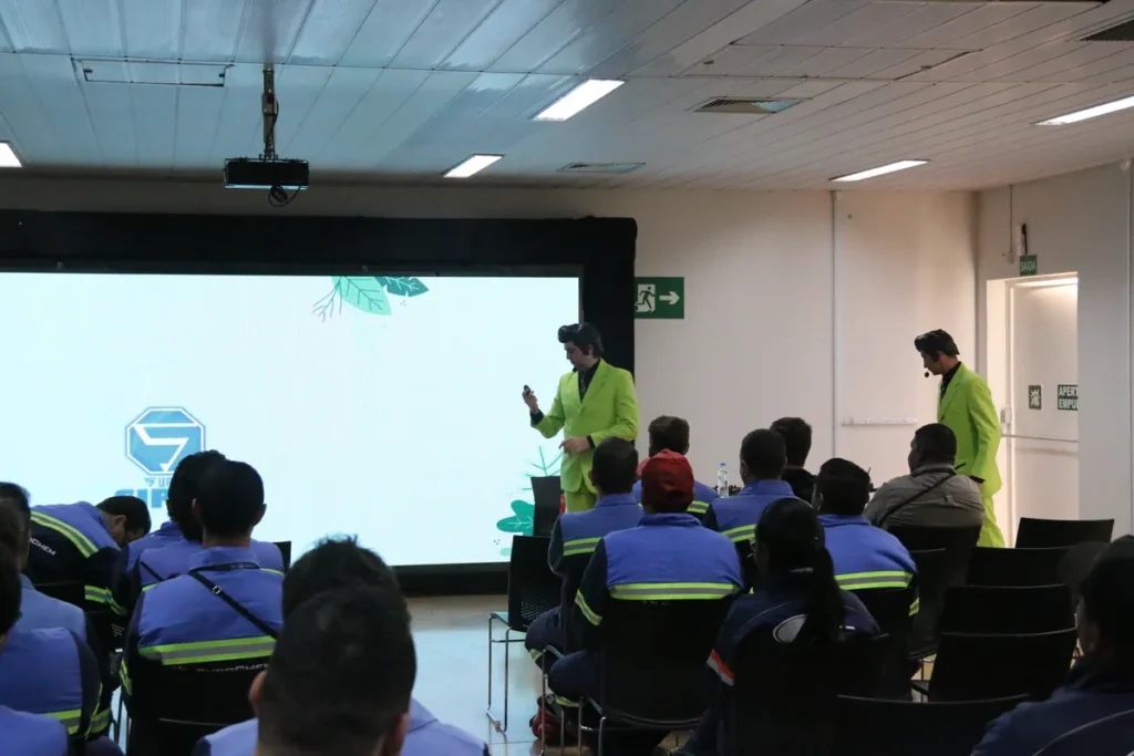 Dois palestrantes em ternos verde-limão e luvas brancas, em um palco, apresentam um treinamento para uma audiência sentada e uniformizada, em um evento da SIPAT.