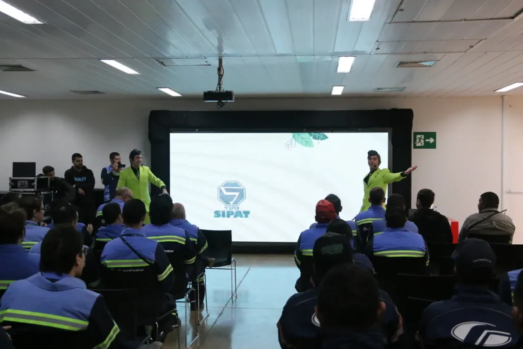 Dois palestrantes em ternos verde-limão e luvas brancas, em um palco, apresentam um treinamento para uma audiência sentada e uniformizada, em um evento da SIPAT.