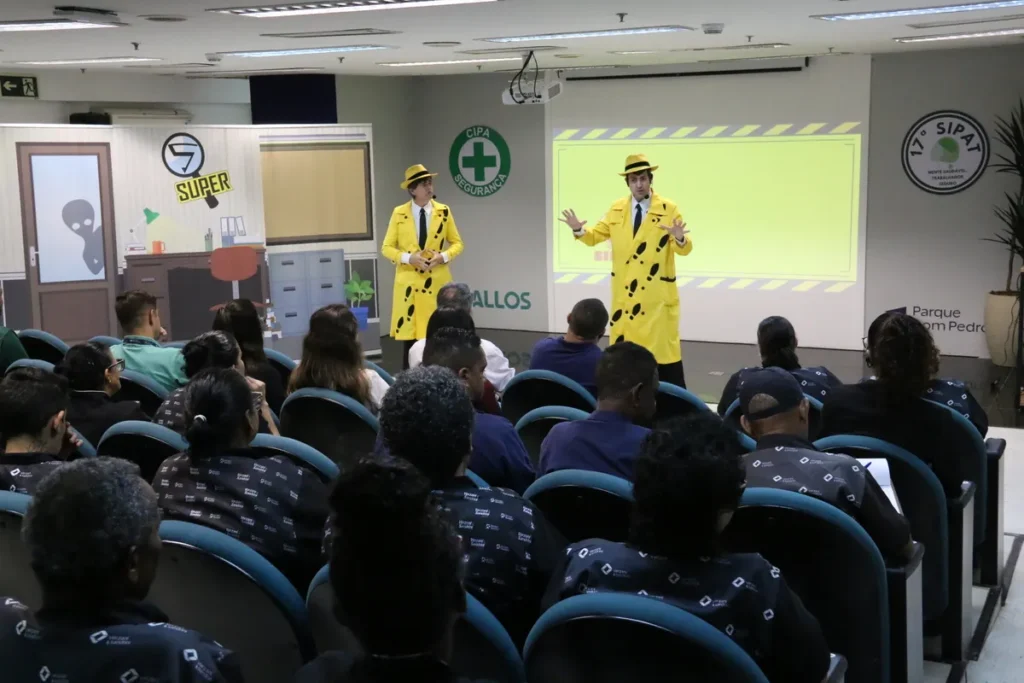 Dois palestrantes vestidos de amarelo e chapéu interagem com a plateia sobre percepção de risco em um evento de segurança corporativo.