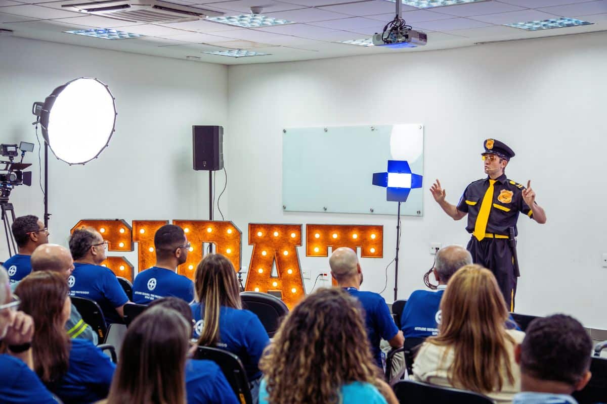 Instrutor ministrando treinamento de percepção de risco para equipe de segurança em sala corporativa equipada com iluminação profissional e painel iluminado.