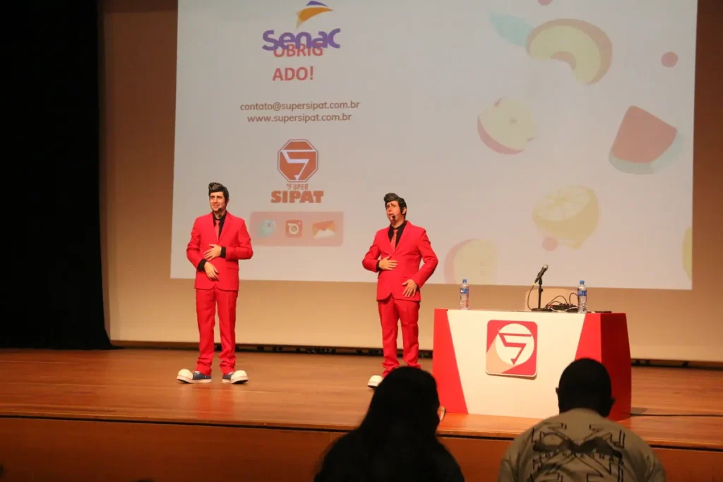 Dupla de apresentadores vestidos de vermelho no palco durante evento de perguntas e respostas para gincana, com telão ao fundo exibindo a logo do Senac e Super Spot.