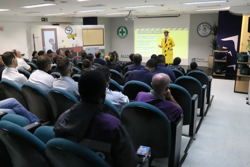 Palestrante fantasiado de amarelo apresenta slides sobre segurança virtual e prevenção de acidentes em um auditório lotado durante a SIPAT.