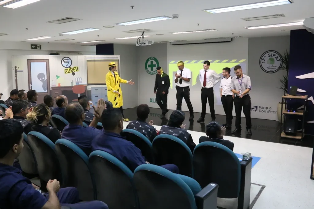 Colaboradores em pé no palco participando de dinâmica de grupo da SIPAT, conduzida por palestrante vestido de amarelo.