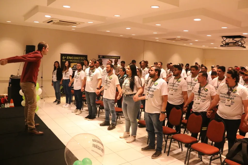 Um grupo de pessoas, vestindo camisetas brancas, estão de pé e com os braços estendidos para os lados, como em uma demonstração ou alongamento. A imagem pode ilustrar uma palestra sobre saúde e bem-estar, com foco na importância do uso de protetor facial.