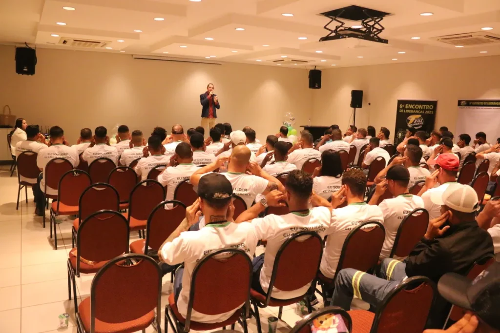 Um homem, que parece ser um palestrante, está em pé em um palco, falando para uma plateia de pessoas sentadas em cadeiras marrons. A imagem sugere uma palestra sobre proteção da pele e uso de protetor facial.