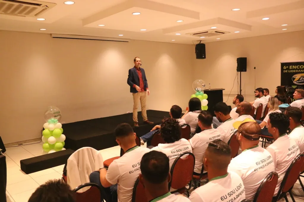 Um homem, que parece ser um palestrante, está em pé em um palco, falando para uma plateia de pessoas sentadas em cadeiras marrons. A imagem sugere uma palestra sobre proteção da pele e uso de protetor facial.