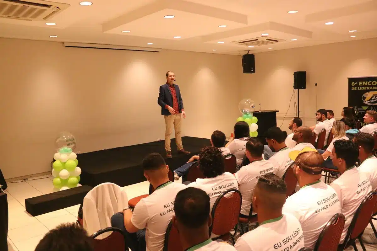 Um homem, que parece ser um palestrante, está em pé em um palco, falando para uma plateia de pessoas sentadas em cadeiras marrons. A imagem sugere uma palestra sobre proteção da pele e uso de protetor facial.