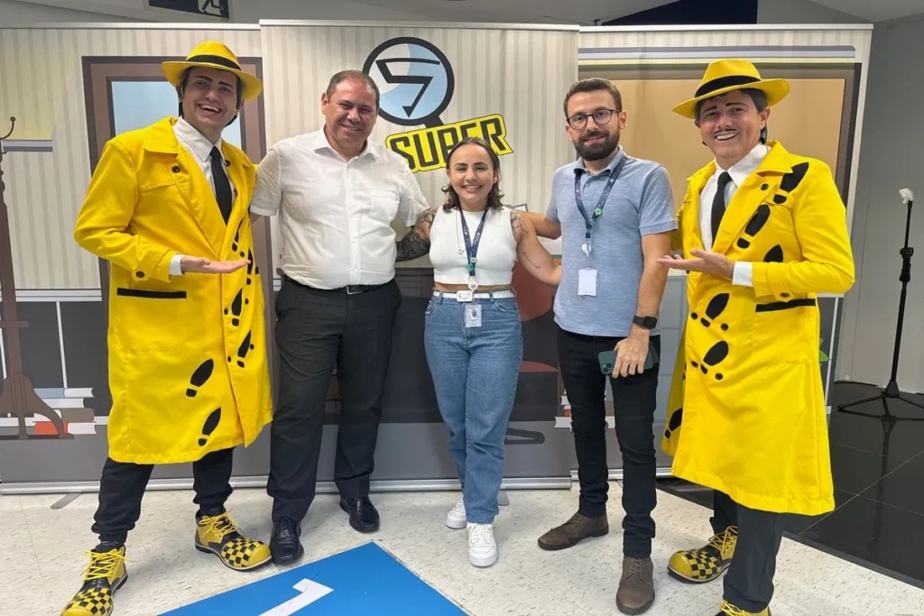 Um grupo de cinco pessoas, três homens e duas mulheres, estão sorrindo para a câmera. Dois dos homens estão vestidos como detetives, com casacos de chuva amarelos e chapéus da mesma cor, enquanto os outros três usam roupas casuais. A imagem representa um evento sobre saúde, focado no protocolo de dor torácica.