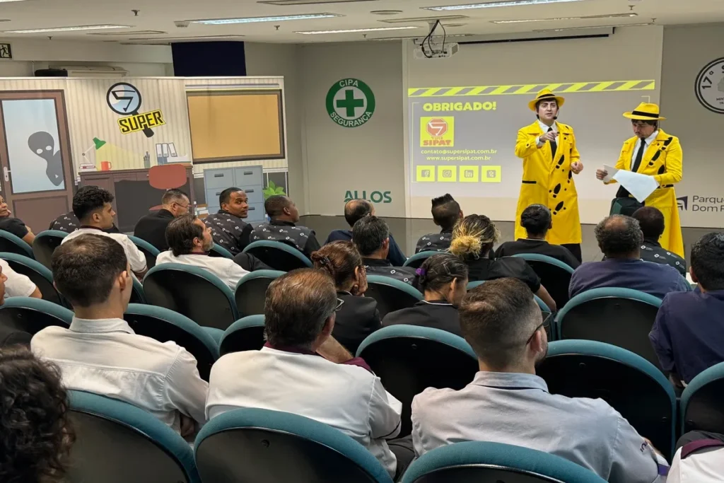 Uma plateia, vista de costas, está assistindo a uma palestra. No telão, dois homens vestidos com casacos de chuva amarelos e chapéus da mesma cor estão em pé, com um logo e um texto de agradecimento ao fundo. A imagem representa o tema de protocolo de dor torácica.