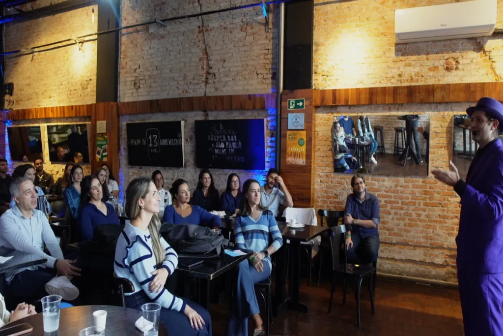 Palestrante de terno roxo gesticulando para um público sentado em mesas em um ambiente rústico com tijolos expostos, em um evento de treinamento.