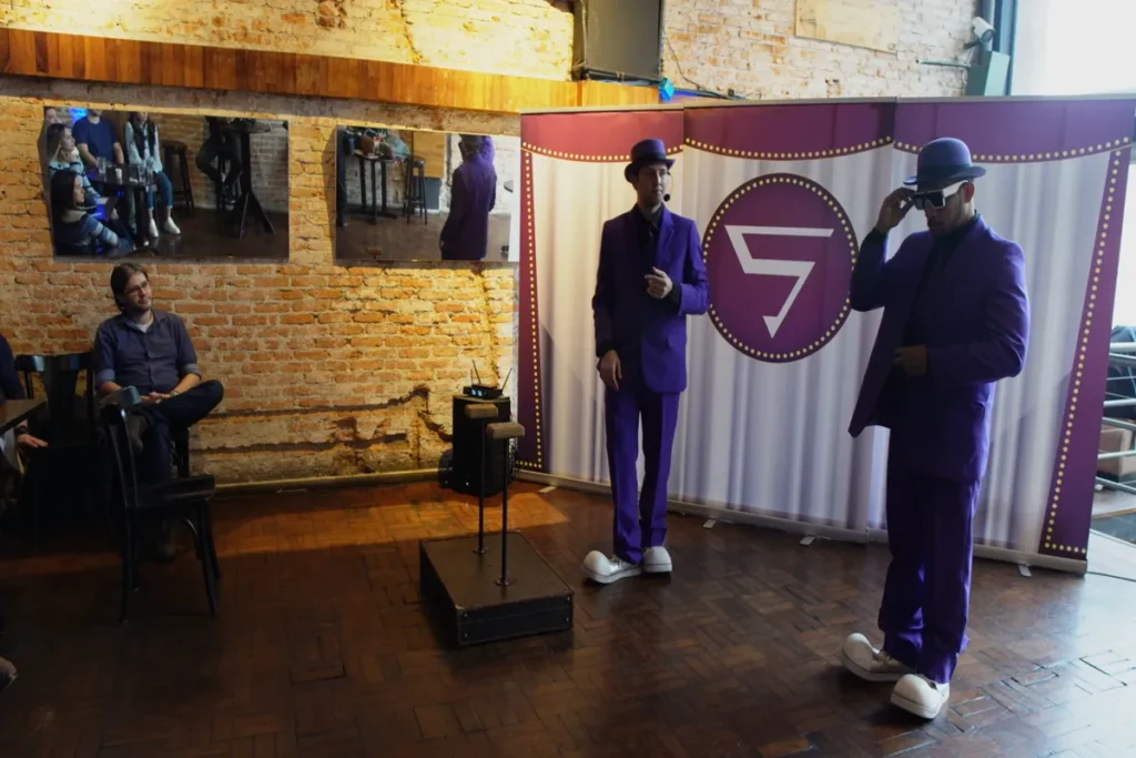 Dois homens vestidos com ternos roxos, chapéus e sapatos grandes, em frente a um banner com o número 5, em um palco de tijolos e madeira, possivelmente em um evento sobre NR 5.