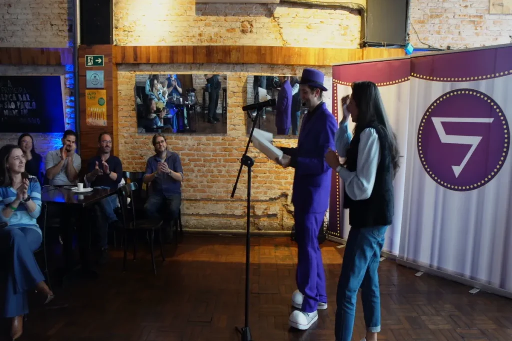 Orador de terno roxo e chapéu entregando um prêmio ou certificado a uma mulher no palco, em frente a um banner do número 5, durante um evento de treinamento.