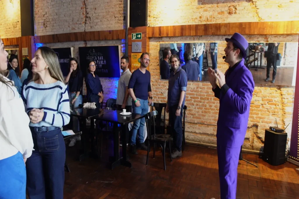 Orador de terno roxo e chapéu interagindo com um pequeno grupo de pessoas em pé em um evento de treinamento em ambiente rústico com tijolos.