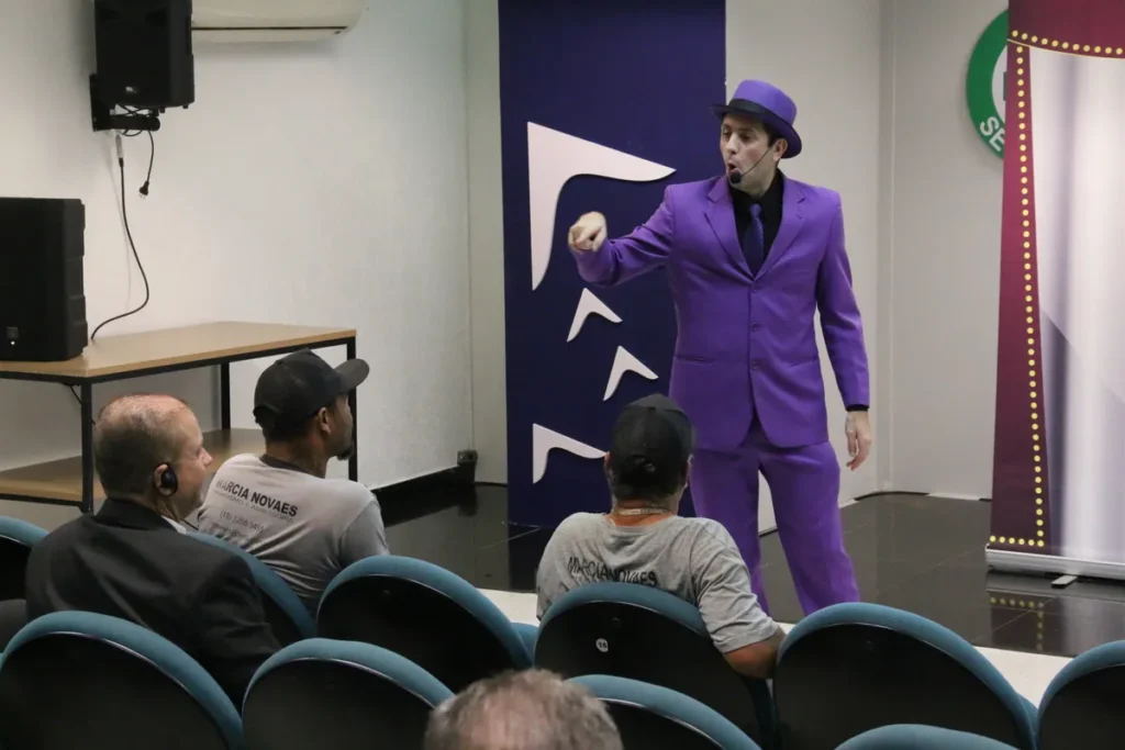 Homem em um palco, com um terno roxo, fazendo um gesto dramático, representando uma encenação de queda da própria altura.