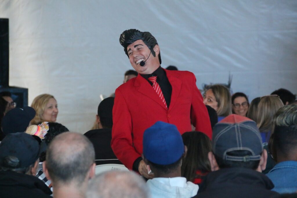 Homem vestido com terno vermelho e microfone headset conduzindo sessão de risoterapia e fazendo o público rir durante evento