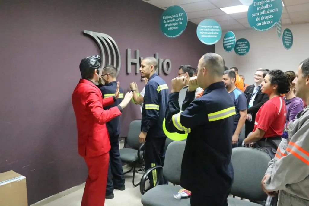 Palestrante de terno vermelho gesticula para um grupo de funcionários (muitos de uniforme azul escuro com faixas refletivas) em uma palestra sobre saúde da mulher no escritório da Hydro.