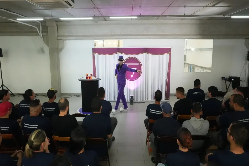 Palestrante de terno roxo e óculos escuros (estilo mágico ou artista) no palco, gesticulando para uma plateia em uma palestra sobre segurança alimentar.