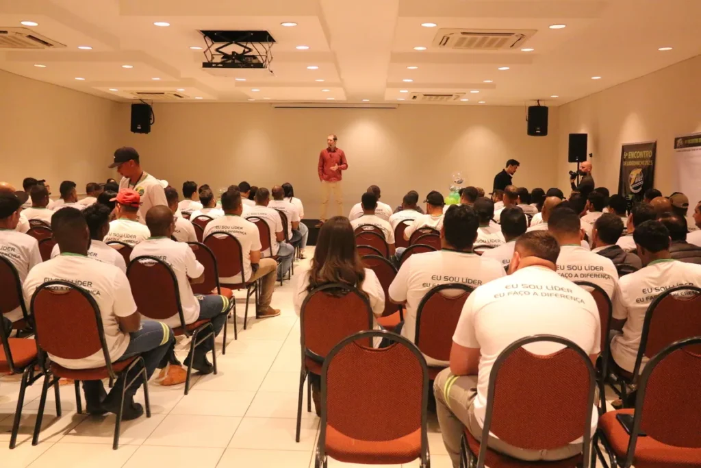 Público de mãos dadas em palestra, com palestrante no palco, em evento de conscientização.