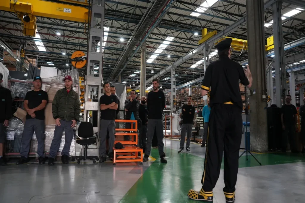 Um instrutor de segurança, vestido de preto e amarelo, está de costas para a câmera, gesticulando para um grupo de trabalhadores em workshop ou treinamento no chão de uma fábrica industrial.