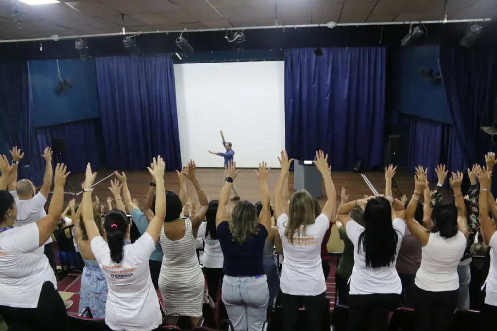 Plateia de auditório com braços levantados em resposta a palestrante, demonstrando engajamento em evento sobre TDAH em adultos ou neurodiversidade.