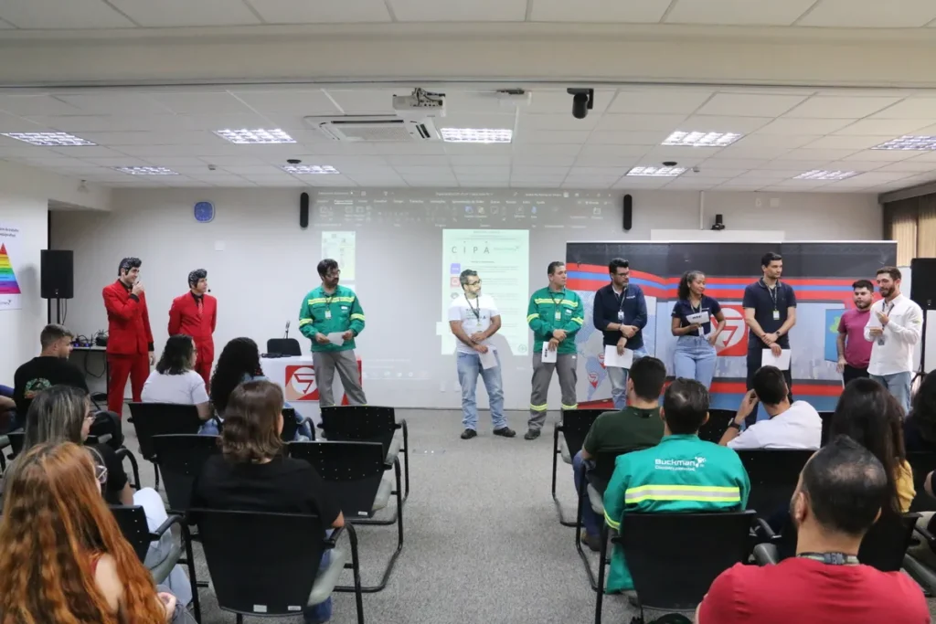 Participantes em pé em um evento de treinamento sobre segurança no trabalho, em um ambiente corporativo. A imagem captura o engajamento do público com os palestrantes, mostrando uma abordagem interativa sobre ergonomia e saúde ocupacional, com foco na atuação do Técnico em Segurança do Trabalho.