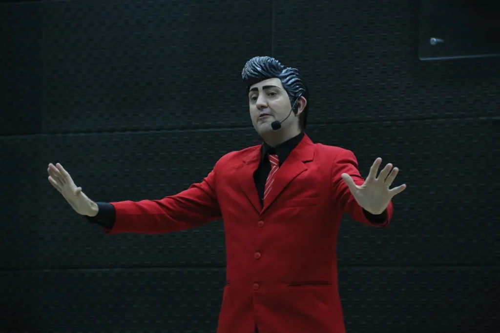 Homem vestido com um terno vermelho vibrante, camisa preta e gravata listrada, com um penteado pompadour preto, gesticulando com as mãos abertas em um palco, ideal para ilustrar apresentações sobre Tendinite e saúde ocupacional.