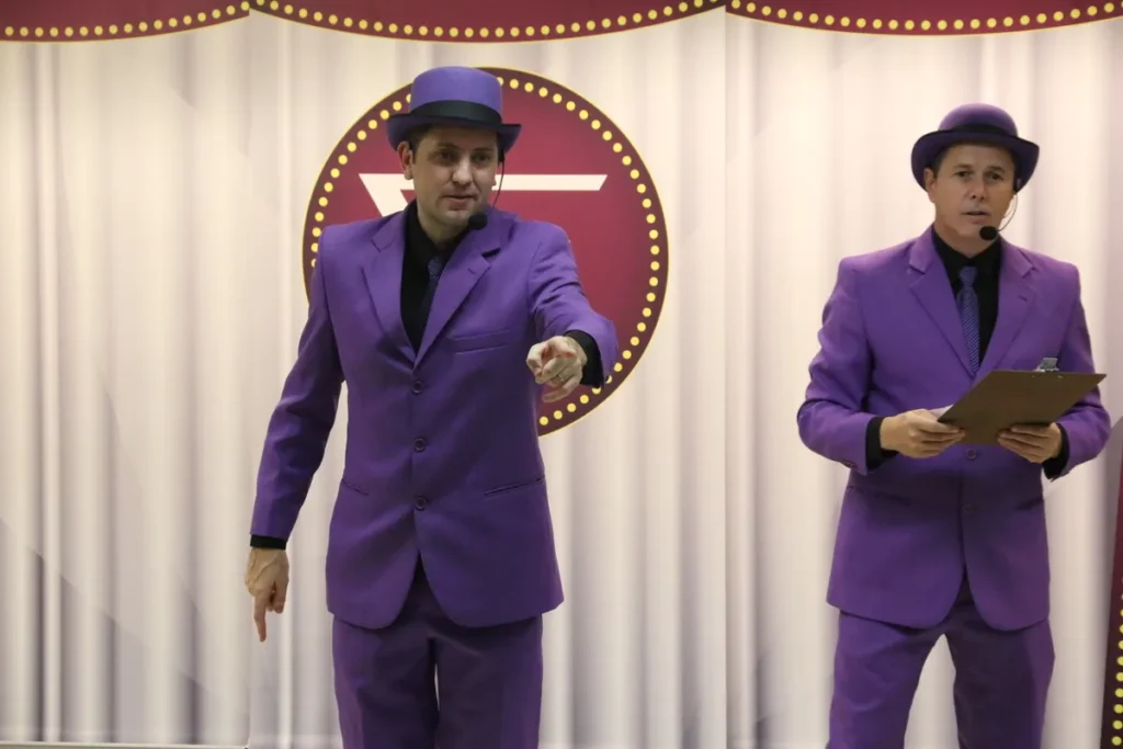 Dois artistas, possivelmente palhaços, vestidos com ternos roxos e chapéus, estão de pé em um palco em frente a uma cortina e um logo. A imagem é de uma apresentação, que pode abordar temas de saúde, como a tosse crônica.