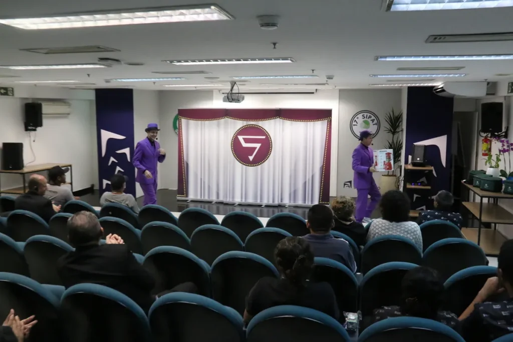Dois palhaços, vestidos com ternos roxos e chapéus da mesma cor, estão em um palco, com uma cortina roxa e branca e um logo no centro. A imagem é de uma palestra ou workshop, que pode abordar temas de saúde, como a tosse crônica.