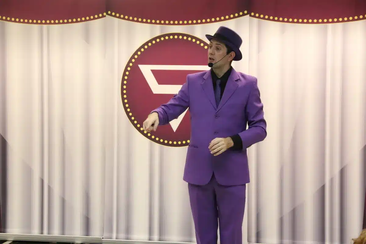Um palhaço, vestido com um terno roxo e chapéu da mesma cor, está em pé em um palco, com uma cortina roxa e branca e um logo no centro. A imagem é de uma palestra ou workshop, que pode abordar temas de saúde, como a tosse crônica.