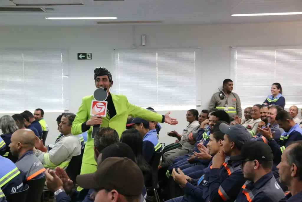 Palestrante carismático com terno verde-limão e microfone em uma palestra motivacional de segurança do trabalho NR 35 Trabalho em Altura, com a plateia de funcionários uniformizados aplaudindo e engajados.