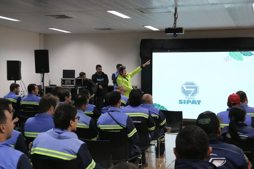 Palestra de Trabalho em Altura - NR 35 durante evento SIPAT. Um instrutor de colete verde aponta para uma tela com o logotipo SIPAT, enquanto uma plateia de funcionários uniformizados assiste ao treinamento de segurança.