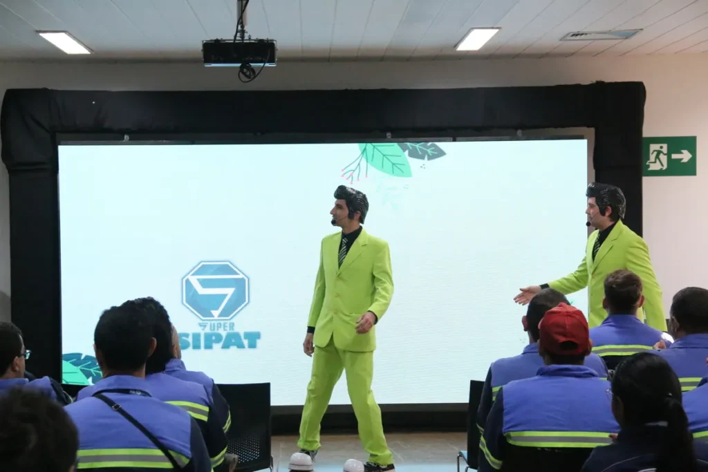 Dois palestrantes idênticos em ternos verde-limão e penteados chamativos em frente a um telão com o logotipo "SUPER SIPAT". Eles conduzem um treinamento dinâmico e motivacional sobre a NR 35 Trabalho em Altura para funcionários uniformizados.