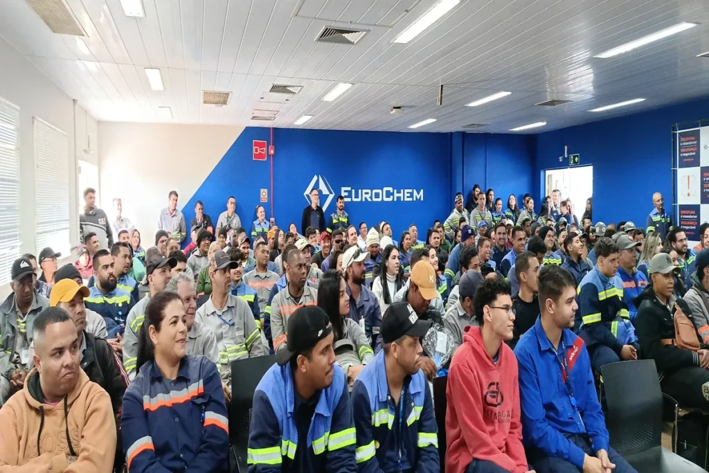 Multidão de funcionários, muitos vestindo uniformes de trabalho, sentados em um auditório azul, assistindo atentamente a uma palestra.