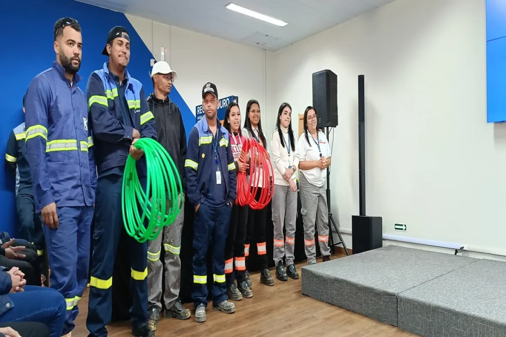 Grupo de colaboradores de uma empresa, alguns com uniformes e segurando cabos e mangueiras, em pé em um palco, participando de uma palestra sobre ultrassonografia abdominal total.